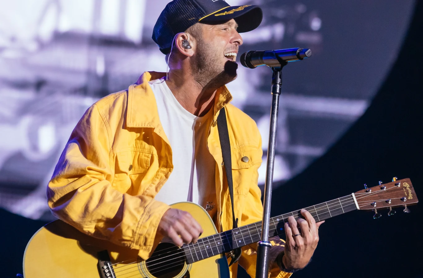 OneRepublic zagra w Krakowie 30 czerwca Mężczyzna w żółtej kurtce i czapce z daszkiem śpiewa do mikrofonu, grając na gitarze podczas koncertu na scenie oświetlonej światłem scenicznym.