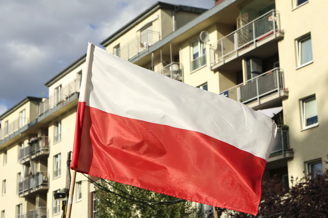 Flaga Polski Polska flaga powiewająca na tle wielopiętrowego budynku mieszkalnego z balkonami i oknami.