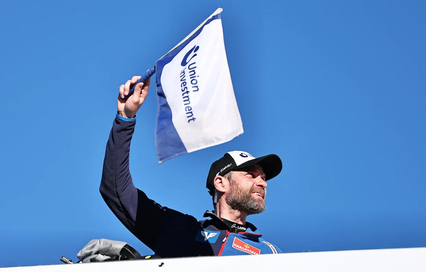 Mężczyzna w sportowej czapce i kurtce unosi flagę z napisem Union Investment na tle jasnoniebieskiego nieba.