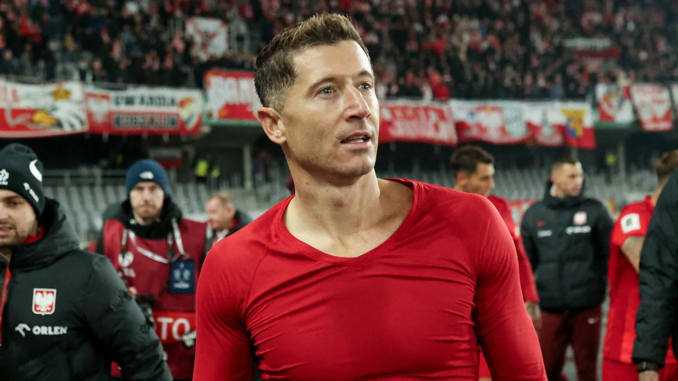 Robert Lewandowski w reprezentacji Polski Piłkarz w czerwonej koszulce znajduje się na stadionie, za nim widoczne są trybuny pełne kibiców z biało-czerwonymi flagami. W tle kilku innych członków drużyny oraz przedstawiciele sztabu, wszyscy skoncentrowani na wydarzeniu sportowym.