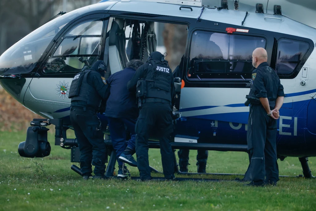 Niemcy. Rusza proces sprawcy ataku na jarmark bożonarodzeniowy w Magdeburgu Grupa funkcjonariuszy w czarnych mundurach eskortuje osobę do policyjnego helikoptera, jeden z funkcjonariuszy stoi z boku obserwując sytuację. Scena rozgrywa się na trawiastej polanie obok lądowiska.