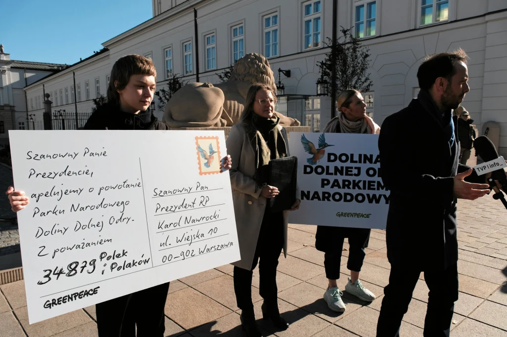 Grupa osób podczas protestu ekologicznego przed budynkiem urzędowym, trzymają transparenty z apelem o utworzenie Parku Narodowego Dolnej Odry oraz dużą symboliczną pocztówkę adresowaną do prezydenta RP. Jedna z osób rozmawia z przedstawicielem mediów, ...