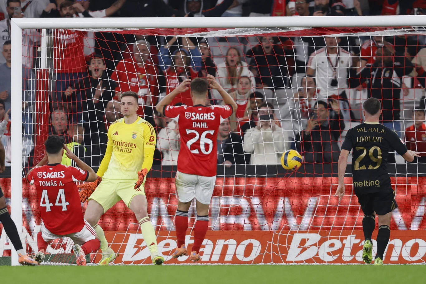 Piłkarze na boisku przy bramce, jeden z nich klęczy, bramkarz i pozostali zawodnicy wyrażają emocje po rozegranej akcji, wokół widoczna publiczność na trybunach.