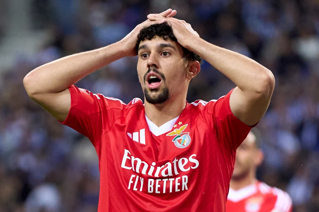 Piłkarz w czerwonej koszulce z herbem Benfiki i napisem 'Emirates Fly Better' trzyma ręce na głowie, wyrażając emocje zaskoczenia lub rozczarowania podczas meczu, w tle rozmyci kibice i zawodnik.