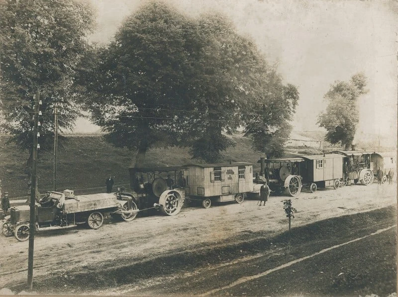 Stare samochody ciężarowe oraz wozy kolejowe zaparkowane na drodze pod dużymi drzewami, kilku mężczyzn stoi wokół pojazdów, scena utrzymana w sepii i stylu typowym dla fotografii z początku XX wieku.