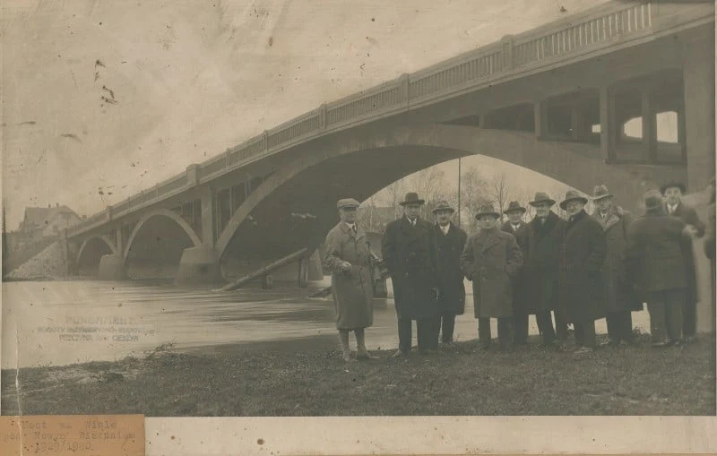Grupa elegancko ubranych osób stojąca na brzegu rzeki w pobliżu dużego mostu łukowego, w tle zabudowa miejska i fragment ulewnego nieba.