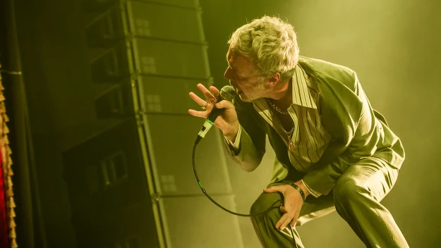 Mężczyzna z mikrofonem w ręku pochylony do przodu podczas śpiewu na scenie, intensywne żółte oświetlenie podkreślające jego sylwetkę, w tle kolumny głośnikowe.