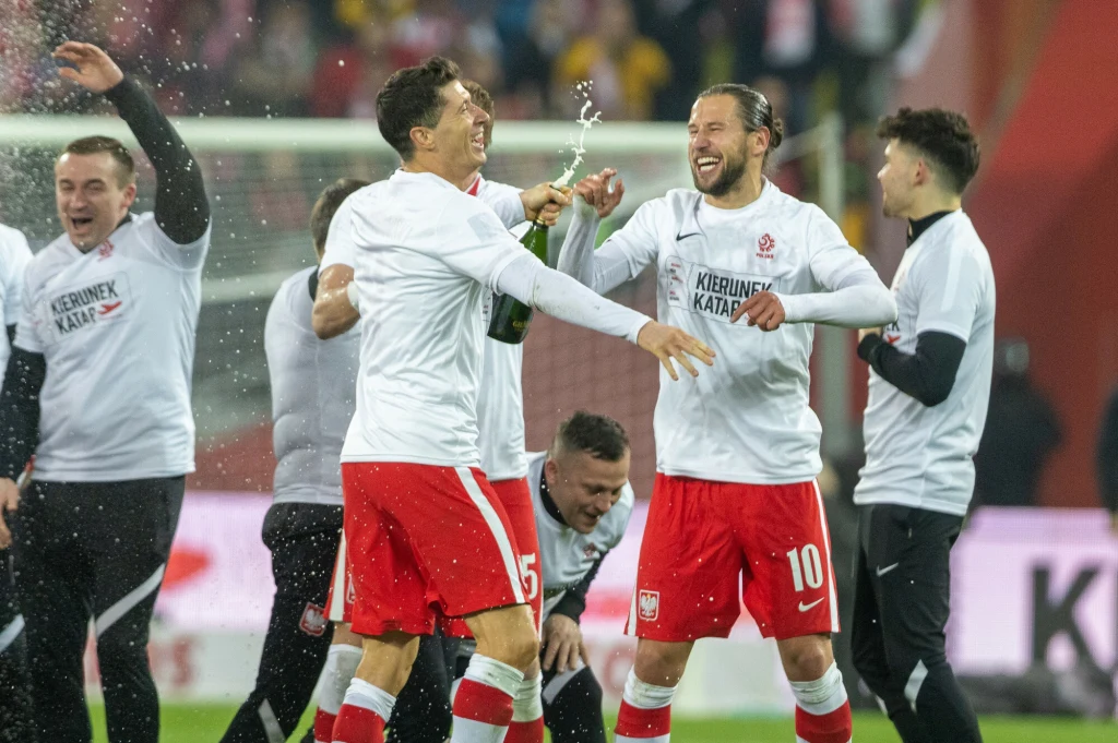 Grzegorz Krychowiak i Robert Lewandowski w reprezentacji Polski Piłkarze w biało-czerwonych strojach świętują zwycięstwo na stadionie, otwierając szampana i okazując radość. Na ich twarzach widoczny szeroki uśmiech, wokół zgromadzeni są inni członkowie drużyny.