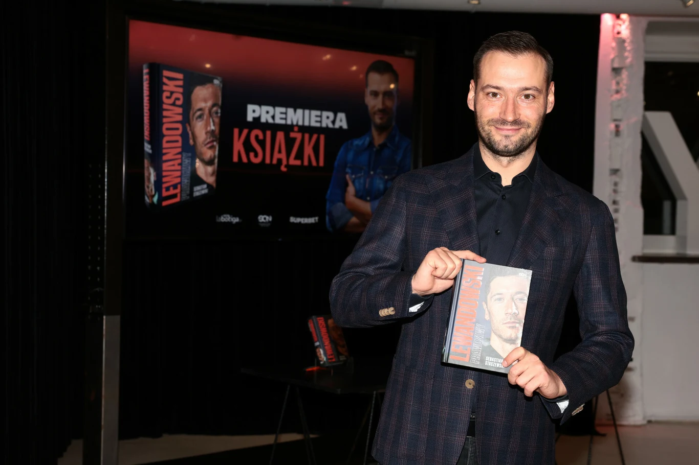 Kadr z premiery książki "Lewandowski. Prawdziwy". Na zdjęciu autor Sebastian Staszewski Mężczyzna ubrany w ciemną marynarkę trzyma w dłoni książkę z wizerunkiem znanego sportowca na okładce, w tle ekran z napisem promującym premierę książki oraz zdjęciem tej samej okładki.