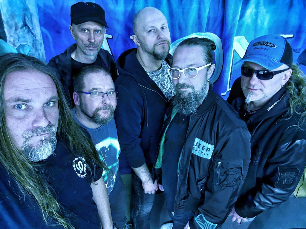 Aion zagra podczas Ostrów Rock Festival 2026 Grupa sześciu mężczyzn ubranych na luzie, częściowo w ciemne kurtki i bluzy, pozuje razem na tle niebieskiego baneru. Jeden z nich nosi czapkę z daszkiem i okulary przeciwsłoneczne, drugi jasne okulary, a pozostali mają zarost lub krótką fryzurę.