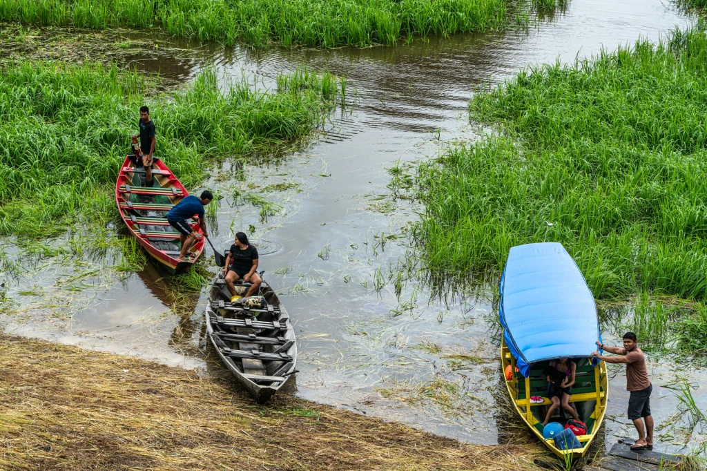Trzy łodzie znajdują się przy brzegu porośniętym zieloną trawą i trzciną. Na łodziach i w ich pobliżu znajduje się kilka osób, z czego dwie osoby stoją na brzegu i przyciągają jedną z łodzi. Jedna z łodzi ma niebieską plandekę, która osłania osoby sied...