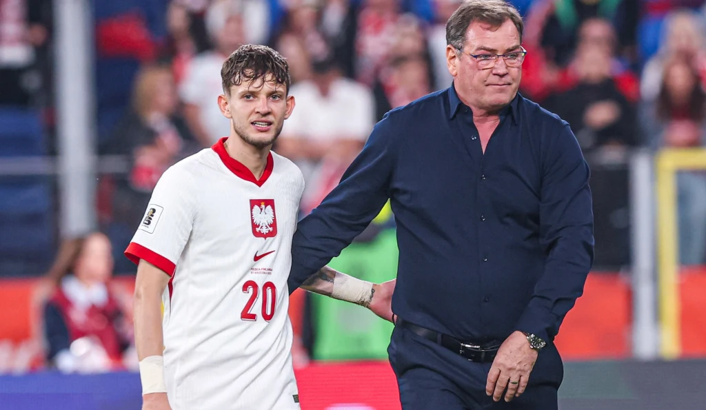 Sebastian Szymański w towarzystwie selekcjonera Jana Urbana Polski piłkarz w białej koszulce z numerem 20 oraz opiekun drużyny w ciemnej koszuli na stadionie, w tle kibice i barwy narodowe.