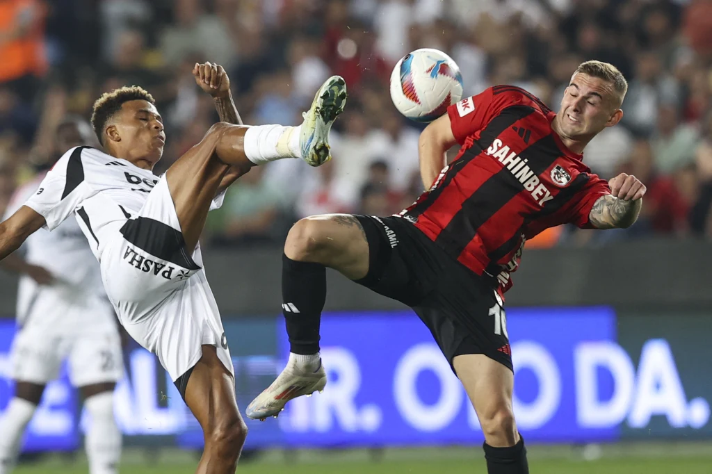 Kacper Kozłowski (z prawej) w barwach Gaziantep FK Dwóch piłkarzy walczy o piłkę w powietrzu na boisku podczas meczu, jeden z nich ubrany w biały strój unosi wysoko nogę, drugi, w czerwono-czarnej koszulce, próbuje przejąć piłkę.