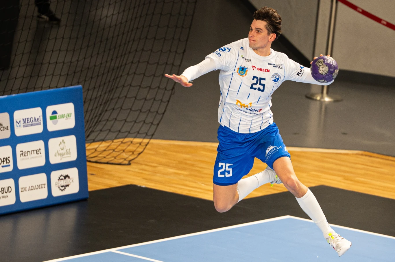 Zawodnik piłki ręcznej w niebieskich spodenkach i białej koszulce z numerem 25 wykonuje dynamiczny skok z piłką nad boiskiem podczas meczu halowego.