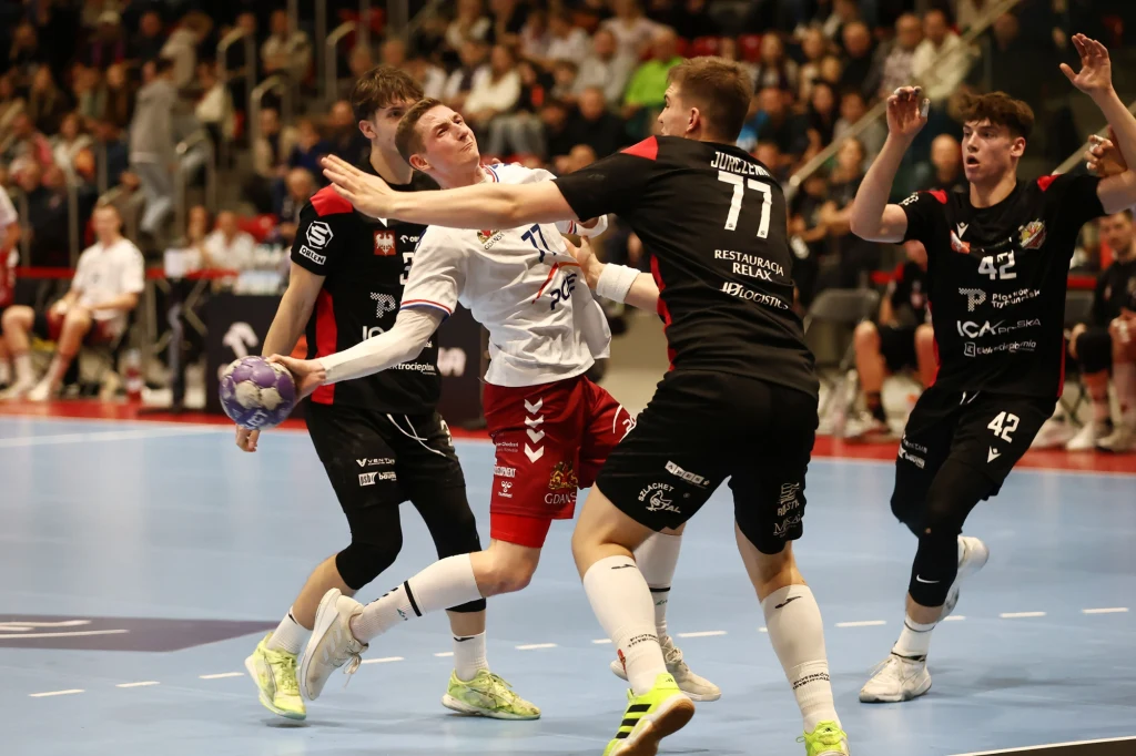 Mecz piłki ręcznej z dynamiczną akcją, zawodnik w białej koszulce próbuje rzucić piłkę, będąc mocno atakowanym przez rywala w czarnej koszulce. W tle widownia skupiona na wydarzeniu sportowym.