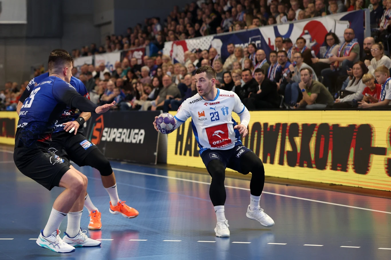 Dwóch zawodników rywalizujących podczas meczu piłki ręcznej na hali sportowej, trybuny wypełnione kibicami, dynamiczna akcja z piłką.