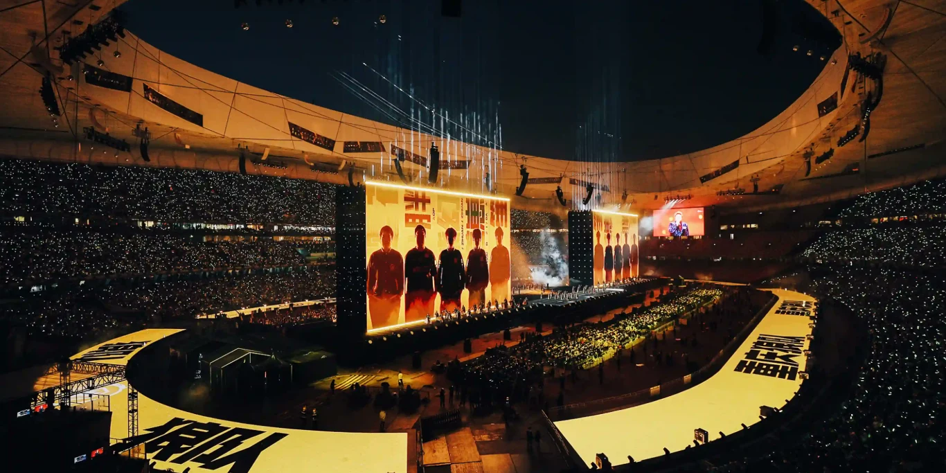 Honor of Kings Tłum ludzi zgromadzony na stadionie, ogromna scena z dużymi ekranami wyświetlającymi sylwetki artystów oraz dynamiczne efekty świetlne tworzące widowiskową atmosferę wydarzenia muzycznego.