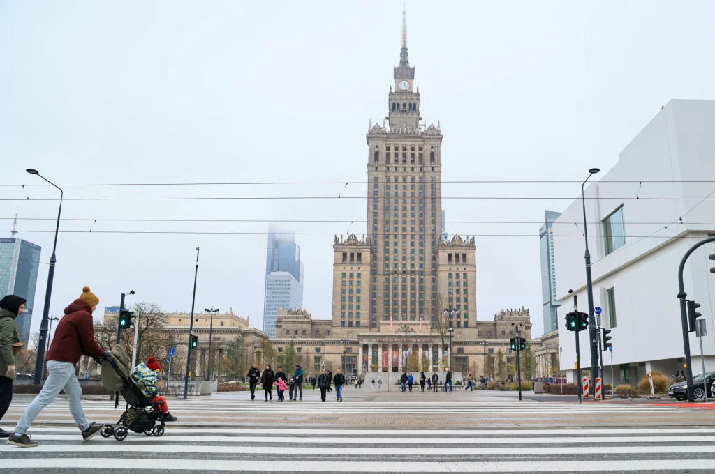 Pałac Kultury i Nauki w Warszawie w centralnej części kadru, otoczony nowoczesnymi budynkami. Na pierwszym planie przejście dla pieszych, przez które przechodzą ludzie, w tym kobieta z wózkiem dziecięcym. Widoczne miejskie latarnie, sygnalizatory świet...