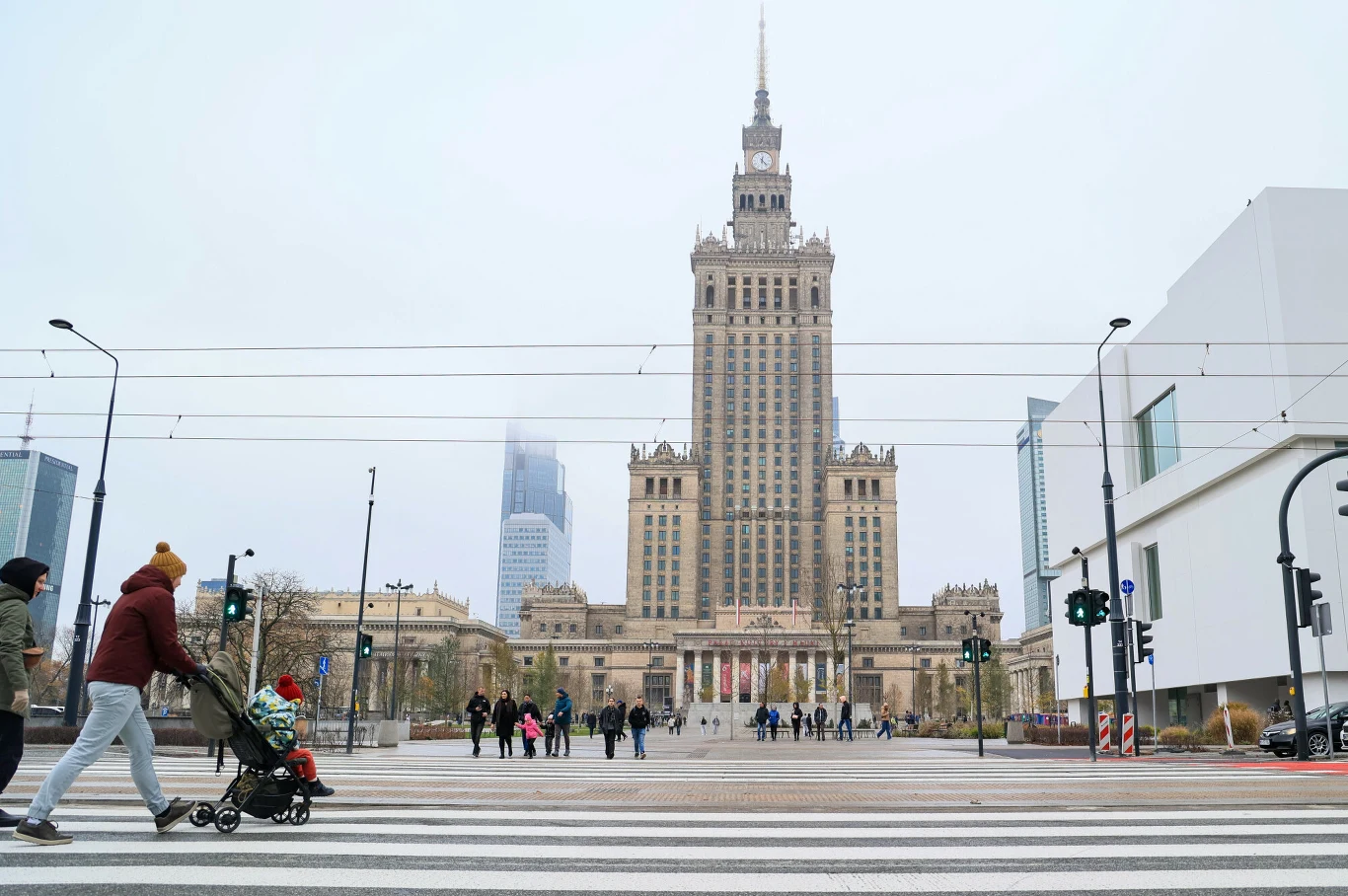 Niemiecki "FAZ" podaje, że Polska może wyprzedzić Francję w obrocie handlowym z Niemcami. Pałac Kultury i Nauki w Warszawie w centralnej części kadru, otoczony nowoczesnymi budynkami. Na pierwszym planie przejście dla pieszych, przez które przechodzą ludzie, w tym kobieta z wózkiem dziecięcym. Widoczne miejskie latarnie, sygnalizatory świet...