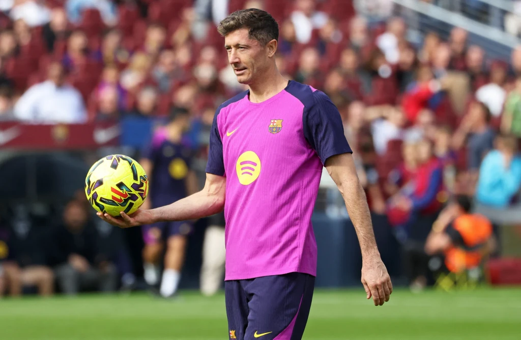 Robert Lewandowski Piłkarz w treningowej koszulce FC Barcelony trzyma piłkę do nogi na tle rozmytego tłumu na trybunach. Wyraz twarzy skupiony, ubranie w klubowe barwy, na przodzie widoczny logotyp sponsora Spotify.