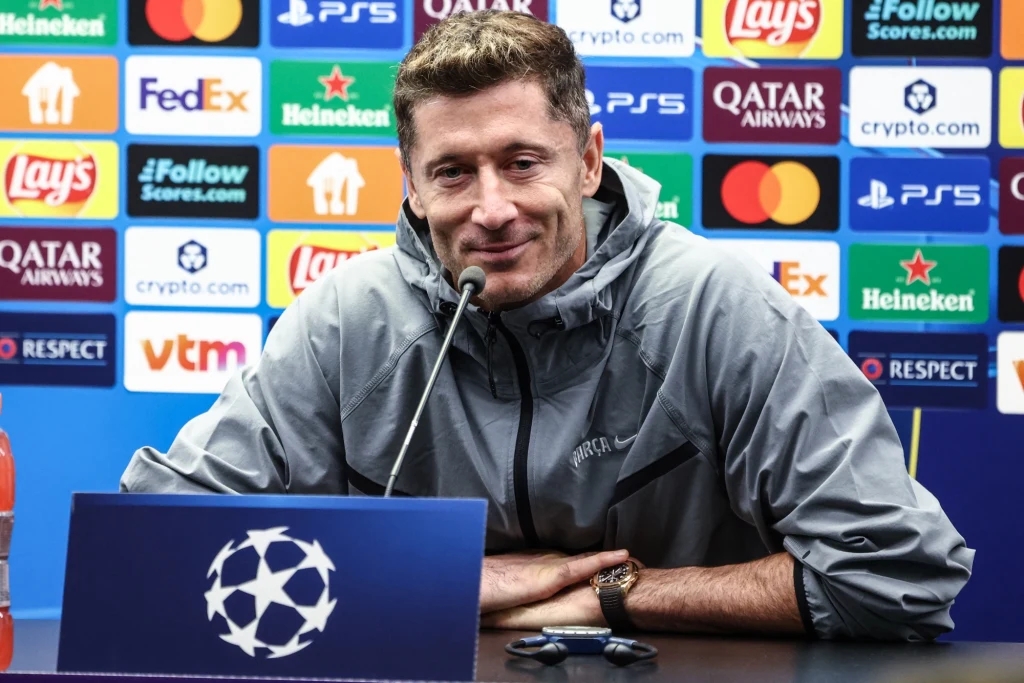 Robert Lewandowski Piłkarz siedzący przy stole konferencyjnym, ubrany w sportową bluzę, z mikrofonem przed sobą. W tle kolorowa ściana sponsorów z widocznymi logotypami znanych marek sportowych i spożywczych.