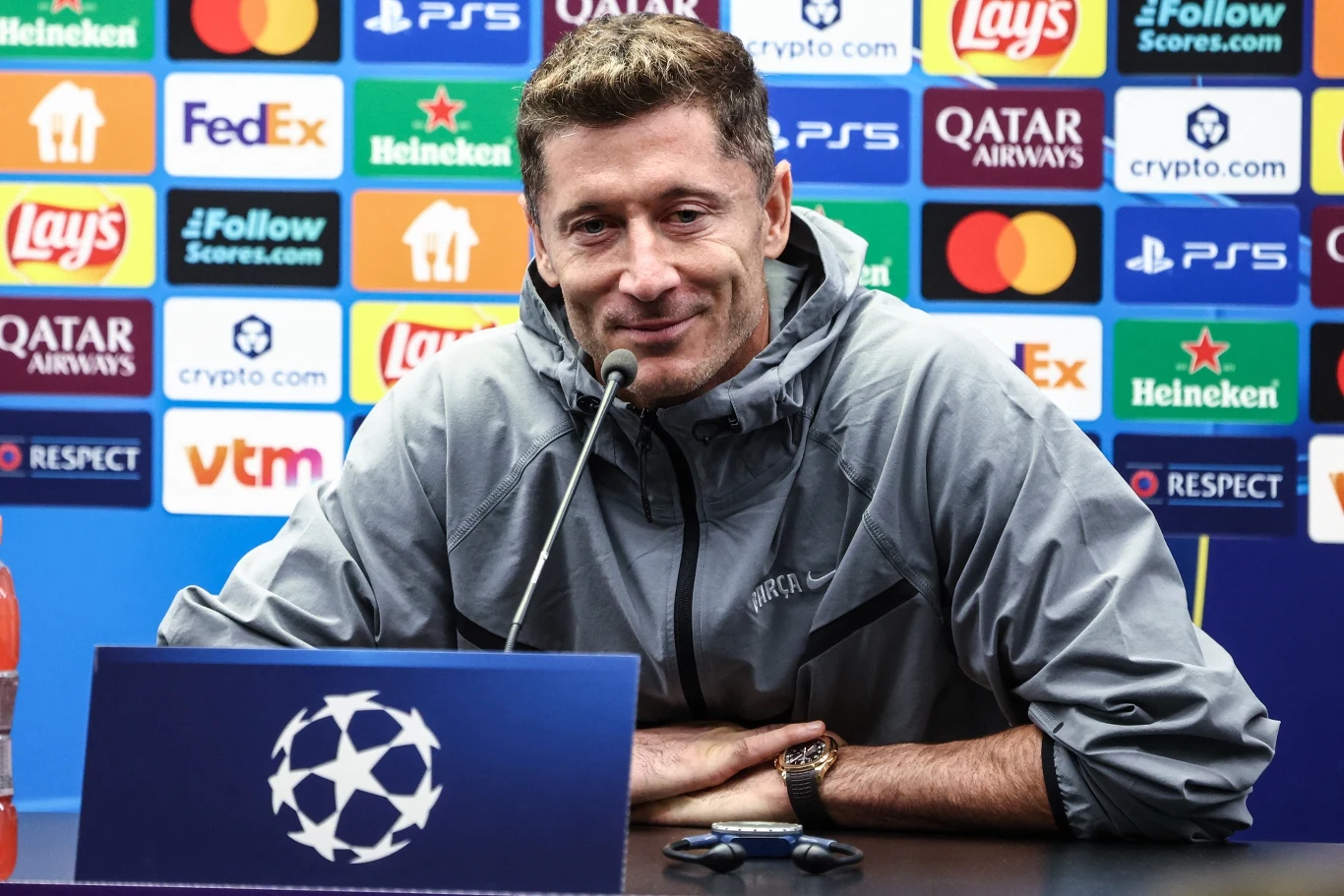 Robert Lewandowski Piłkarz siedzący przy stole konferencyjnym, ubrany w sportową bluzę, z mikrofonem przed sobą. W tle kolorowa ściana sponsorów z widocznymi logotypami znanych marek sportowych i spożywczych.
