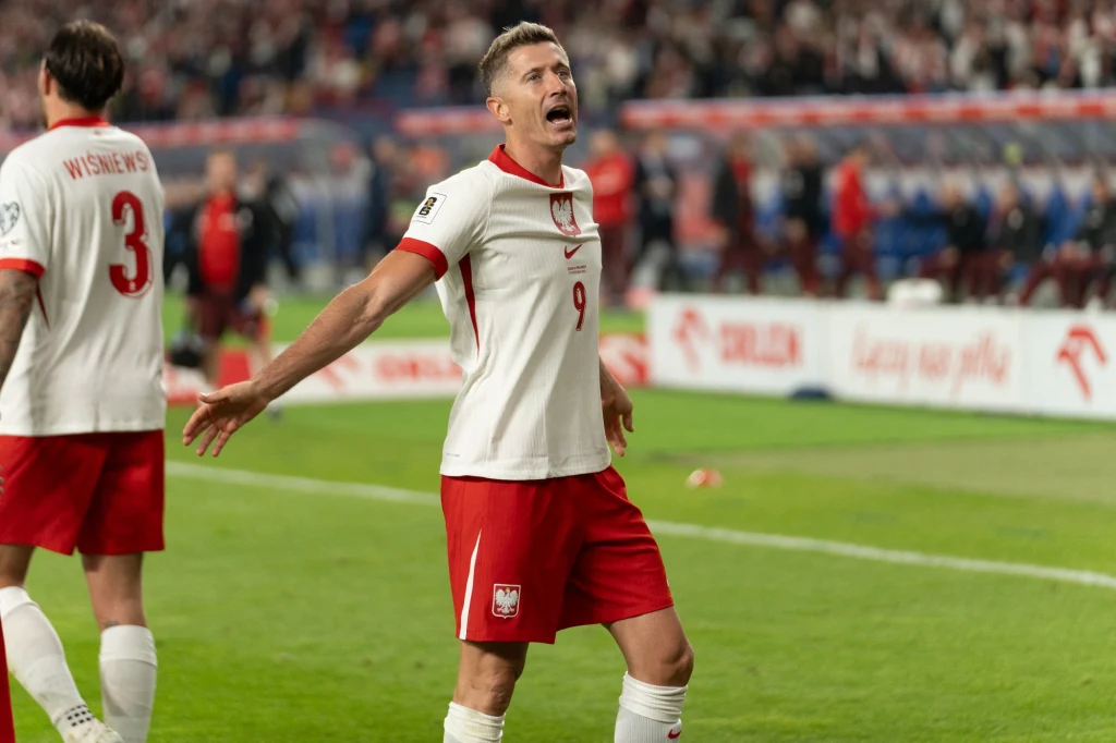 Robert Lewandowski w barwach reprezentacji Polski Piłkarz w biało-czerwonym stroju z numerem 9 na tle boiska piłkarskiego i rozmytego tłumu na trybunach, unosi ramiona z otwartymi ustami, sprawiając wrażenie okazywania silnych emocji podczas meczu.