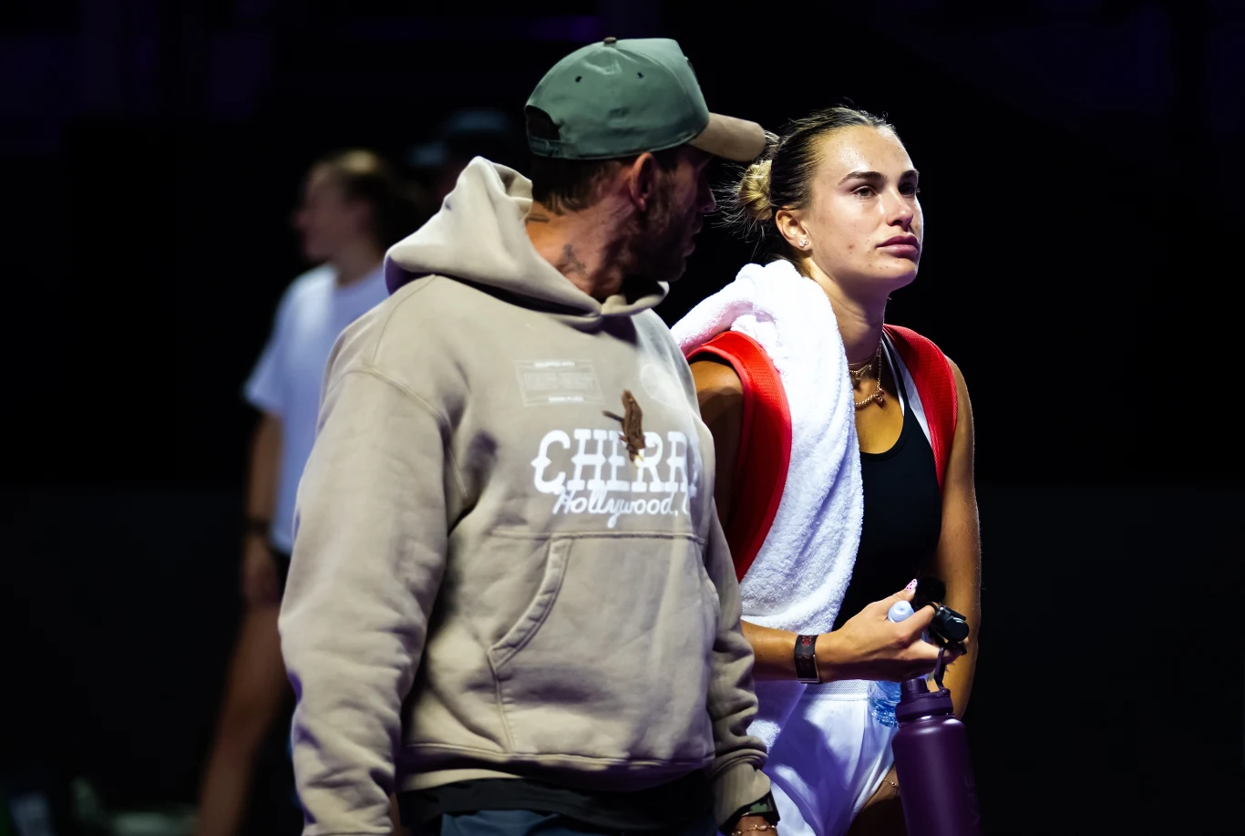 Georgios Frangulis i Aryna Sabalenka Sportowiec z ręcznikiem na ramionach oraz butelką w dłoni stoi obok mężczyzny w bluzie z kapturem i czapce, oboje znajdują się w oświetlonym miejscu z ciemnym tłem, co sugeruje przerwę podczas zawodów sportowych.