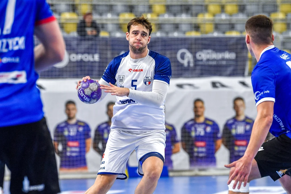 Zawodnik drużyny w białym stroju trzyma piłkę ręczną w dłoni, szykując się do podania lub rzutu, otoczony przez dwóch zawodników drużyny przeciwnej w niebieskich strojach, na tle trybun i banerów reklamowych.