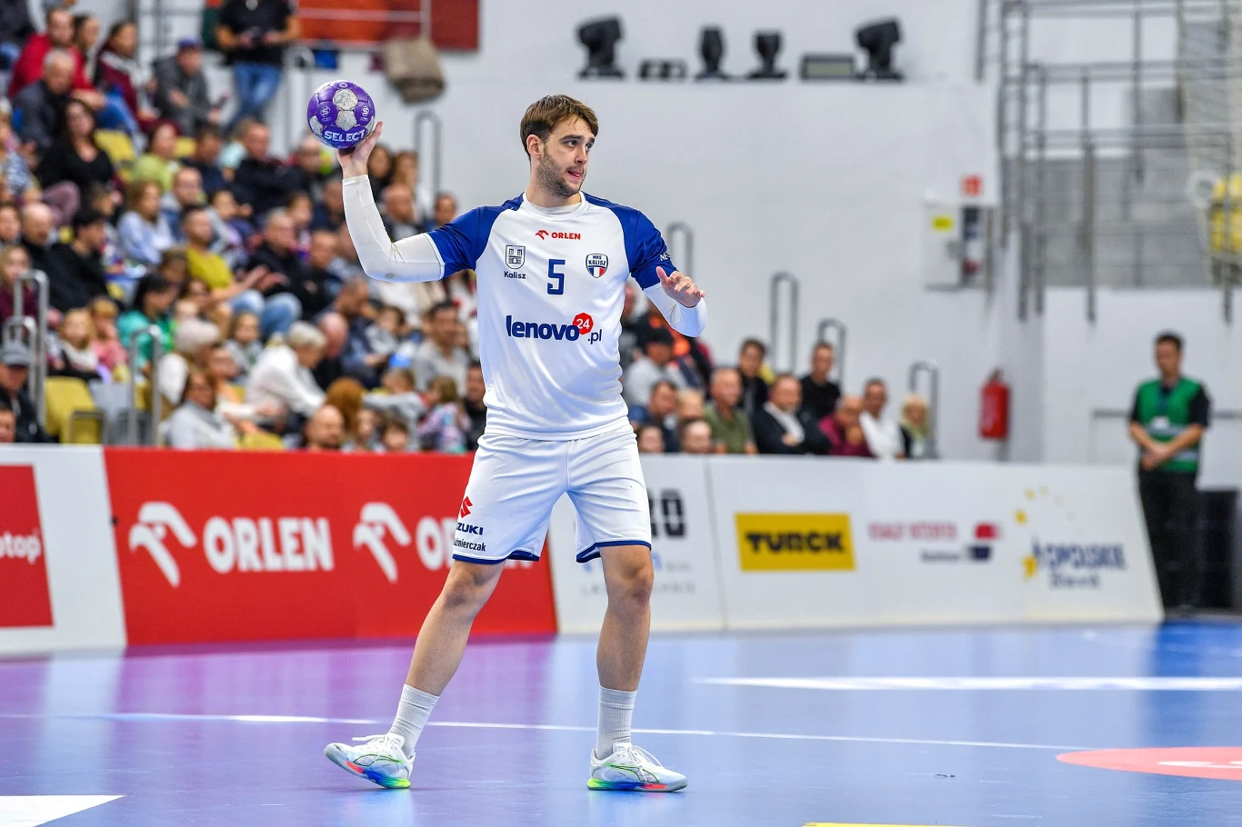 Zawodnik piłki ręcznej w białym stroju z piłką w dłoni gotowy do podania podczas rozgrywki, w tle widoczni kibice na trybunach oraz reklamy na bandach.