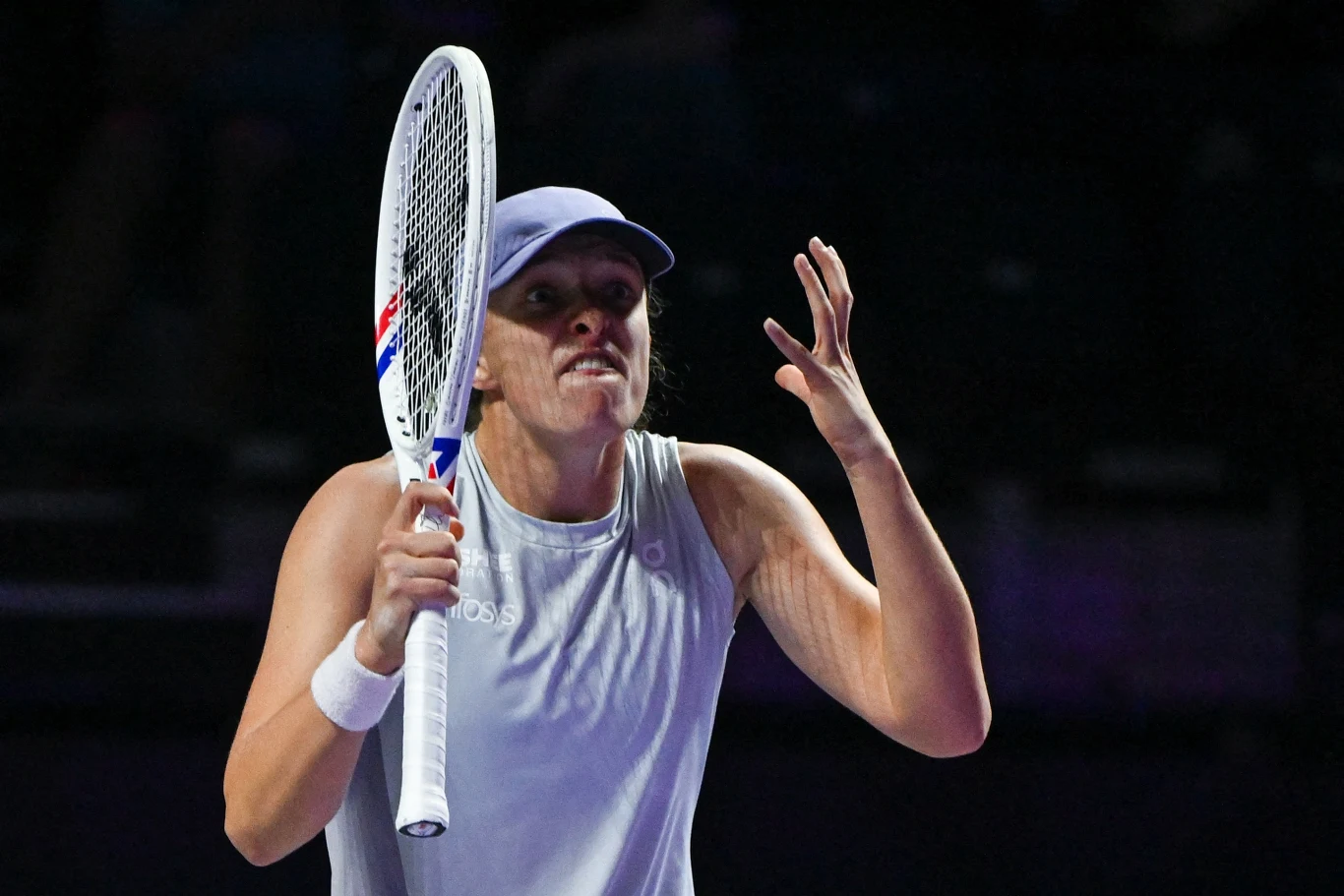 Tenisistka w sportowym stroju i czapce trzyma rakietę tenisową, wykonując energiczny gest ręką oraz wyrażając emocje mimiką twarzy podczas meczu na korcie.