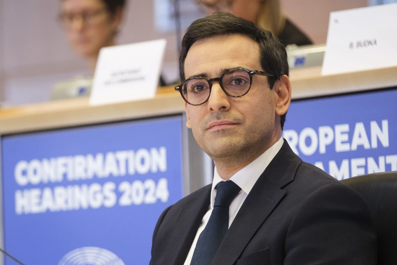 Mężczyzna w garniturze i okularach siedzący przy stole podczas przesłuchań w Parlamencie Europejskim, za nim tablica z napisem Confirmation Hearings 2024 oraz inne osoby w tle.