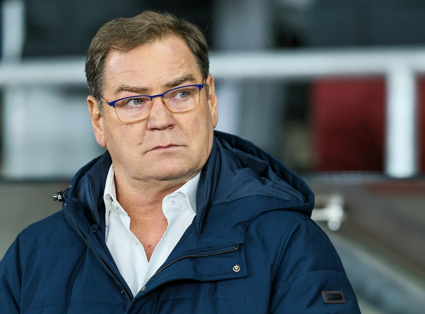 Mężczyzna w okularach oraz granatowej kurtce stoi w tle zamazanego wnętrza, patrząc lekko na bok. Ma poważny wyraz twarzy i starannie ułożone włosy.