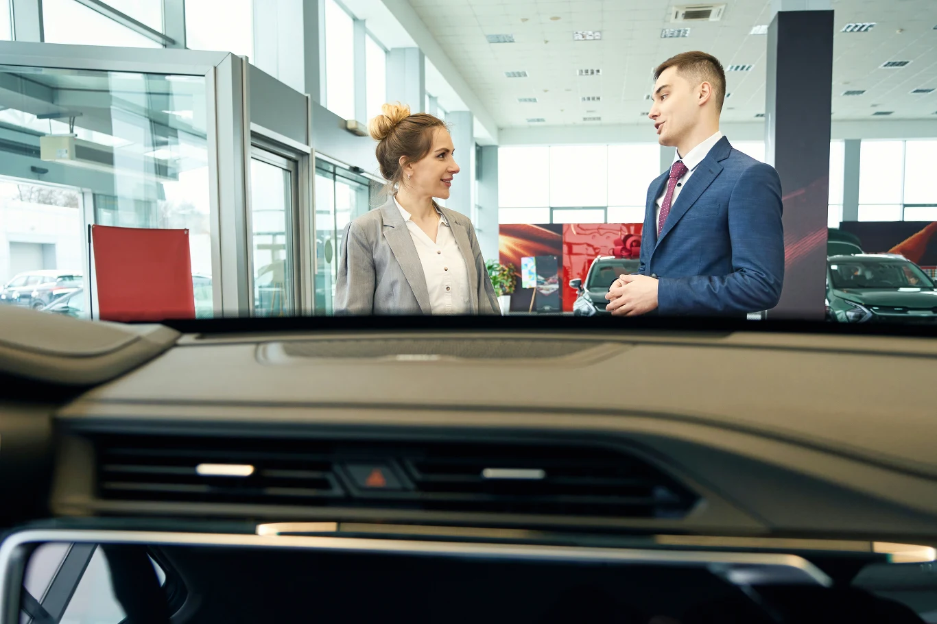 Handlowcy w salonie zaskakują podejściem do klienta. Nie zawsze pozytywnie Dwójka elegancko ubranych osób rozmawia w salonie samochodowym, przez przednią szybę auta widoczną na pierwszym planie widać standy reklamowe oraz nowoczesne wnętrze pełne naturalnego światła.