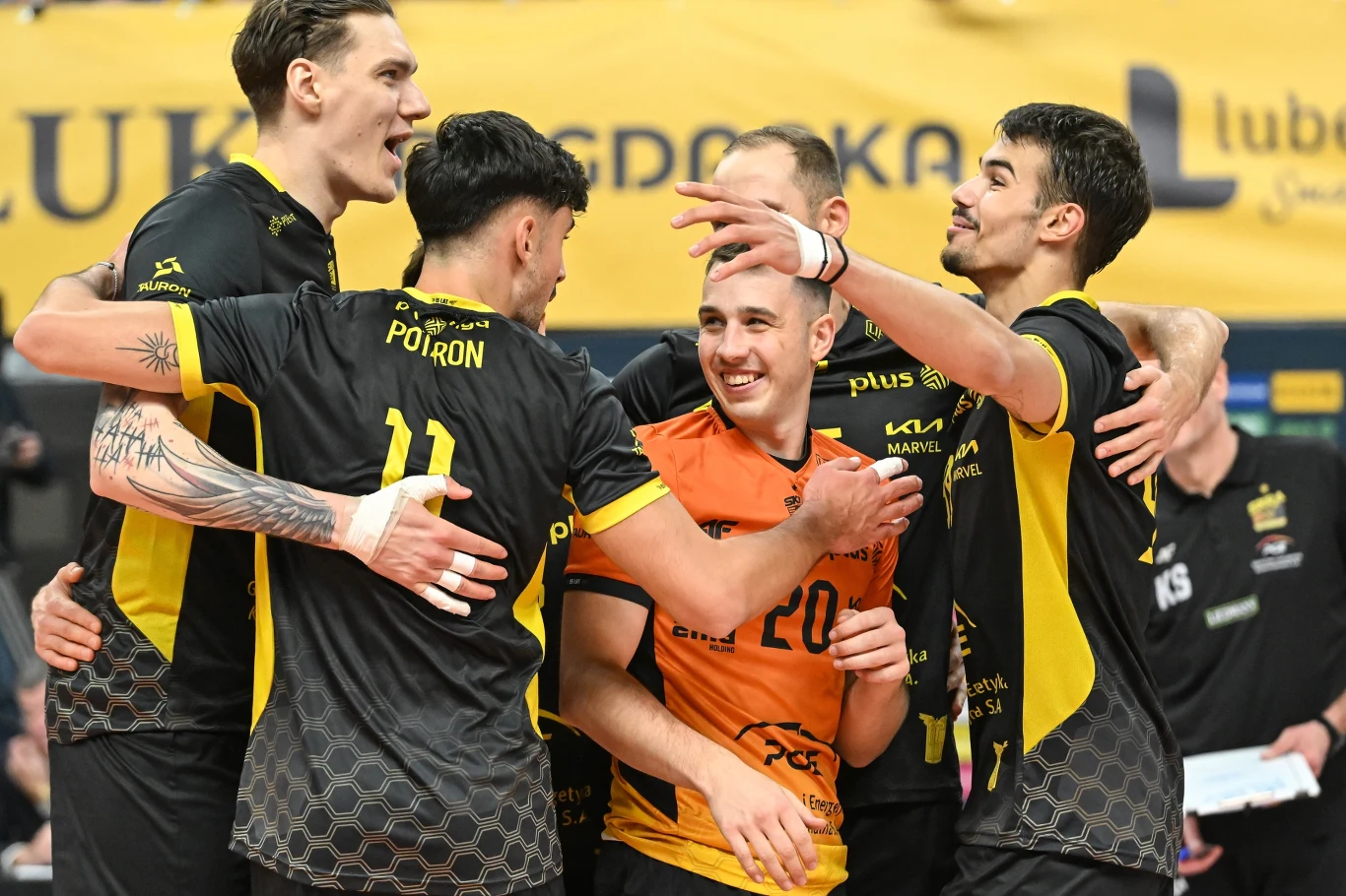 Siatkarze PGE GiEK Skry Bełchatów. Pierwszy z lewej Bartłomiej Lemański Grupa siatkarzy w czarno-żółtych strojach świętuje zdobyty punkt, przytulając się i uśmiechając na tle żółtego banera z napisem.