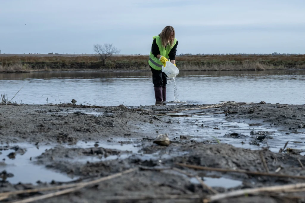 Osoba w żółtej kamizelce odblaskowej zbliża się do brzegu rzeki, trzymając w rękach biały pojemnik, z którego coś wylewa na błotnistą ziemię, otoczona przez surowy, naturalny krajobraz z rzeką i polami w tle.