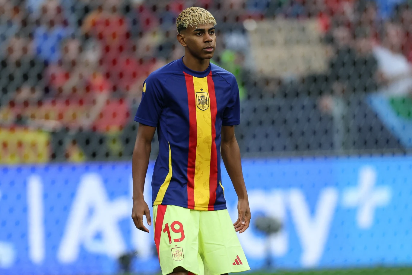 Młody piłkarz w koszulce z numerem 19 w barwach narodowych stoi na murawie stadionu, w tle widoczne rozmyte trybuny z kibicami.