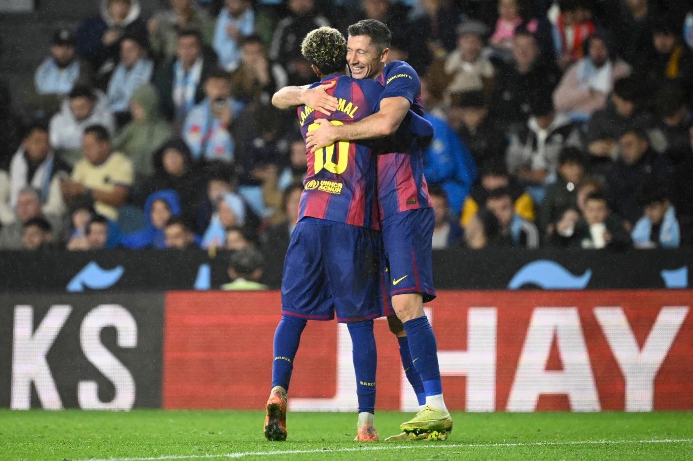 Dwóch piłkarzy w granatowo-bordowych strojach ściska się na murawie stadionu, w tle widoczni kibice na trybunach.