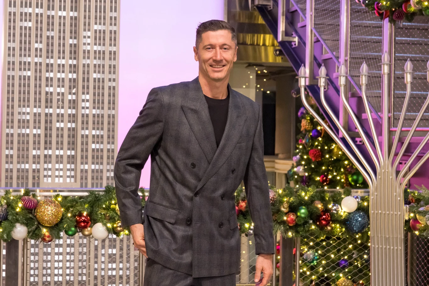 Robert Lewandowski Uśmiechnięty mężczyzna w eleganckim garniturze stoi na tle ozdobionego choinkowymi dekoracjami wnętrza, z widokiem na wysokie budynki i metalowe schody po prawej stronie.