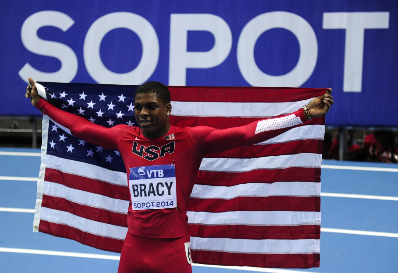 Zawodnik reprezentacji Stanów Zjednoczonych ubrany w strój sportowy trzyma rozpostartą amerykańską flagę na tle niebieskiej bieżni i napisu Sopot. Na piersi widoczny numer startowy z imieniem i nazwiskiem Bracy oraz datą zawodów Sopot 2014.