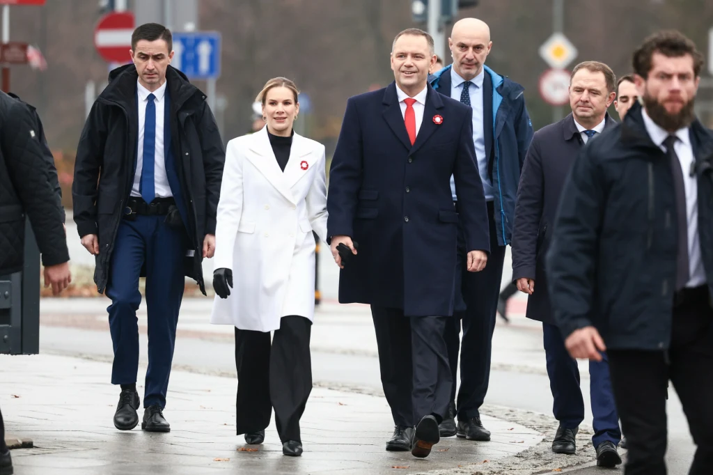 Grupa elegancko ubranych osób w jasnych i ciemnych płaszczach idzie razem po chodniku w przestrzeni miejskiej, w tle znaki drogowe i jesienne drzewa.