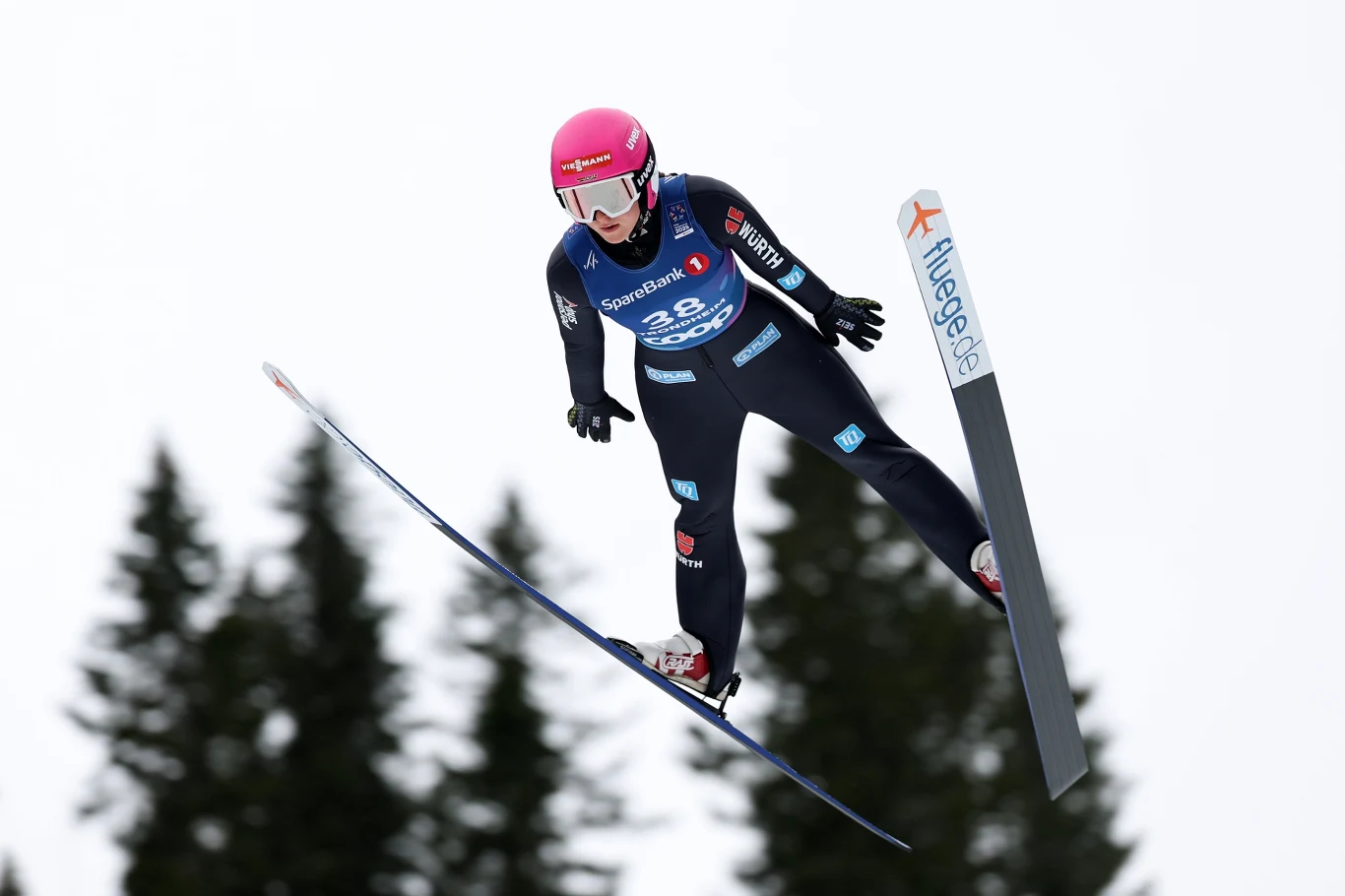 Nathalie Armbruster Zawodniczka w kombinezonie narciarskim w trakcie lotu na skoczni, widoczny narty, kask ochronny oraz numer startowy, w tle rozmazane drzewa i niebo.