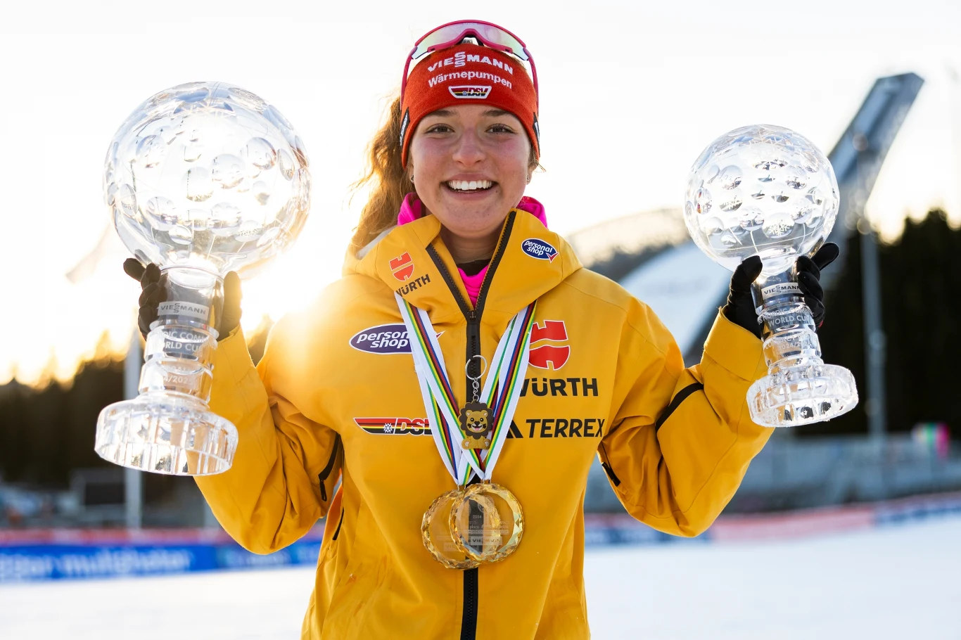 Nathalie Armbruster Młoda sportsmenka w żółtym stroju z medalami na szyi trzyma w obu rękach duże, kryształowe trofea. Uśmiechnięta postać stoi na tle zaśnieżonego stadionu sportowego z widocznymi elementami toru biathlonowego.