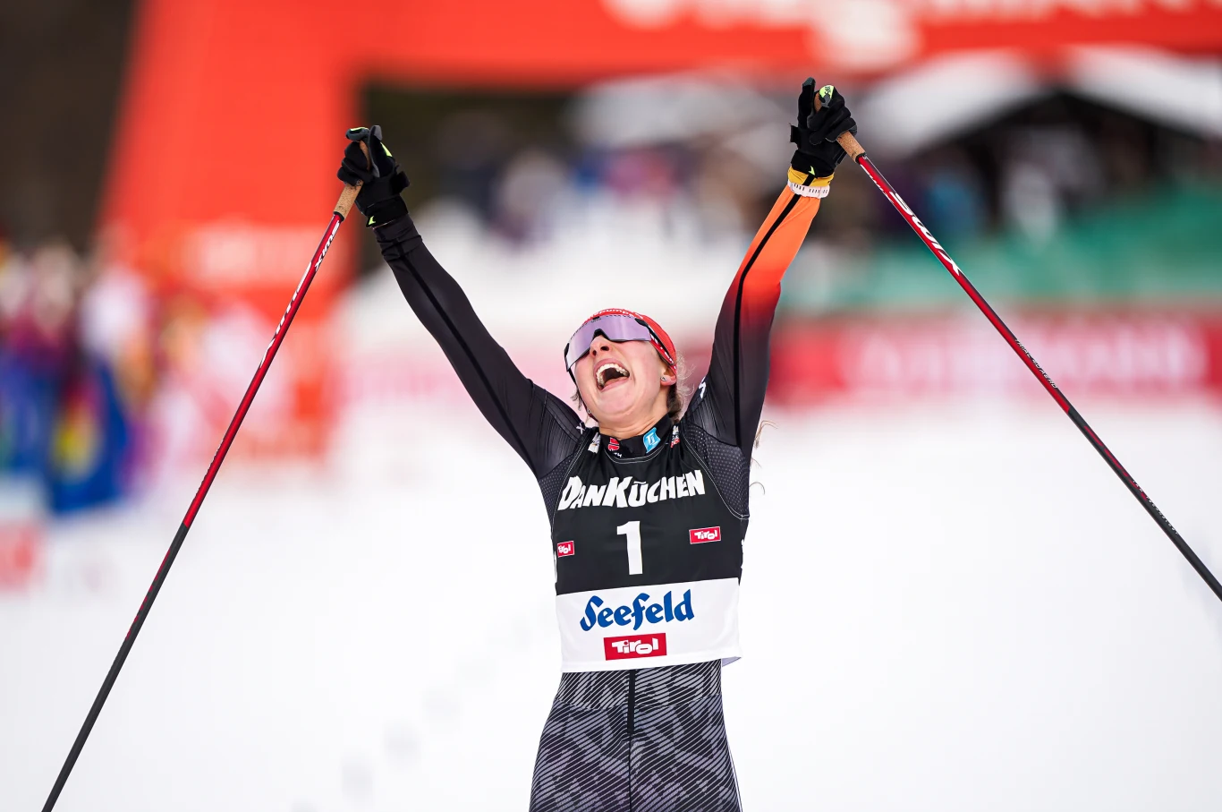 Nathalie Armbruster Narciarka biegowa przekracza linię mety z uniesionymi do góry rękami trzymającymi kijki, wyrażając radość i triumf, na stroju widoczne numery oraz nazwy sponsorów, w tle rozmazana publiczność i elementy dekoracyjne zawodów.