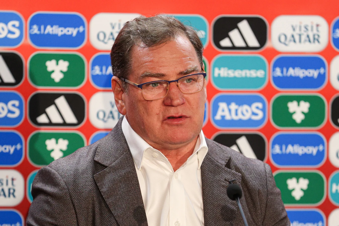 Jan Urban Mężczyzna w okularach i marynarce podczas konferencji prasowej, stoi przed ścianą z wieloma logotypami sponsorów sportowych.