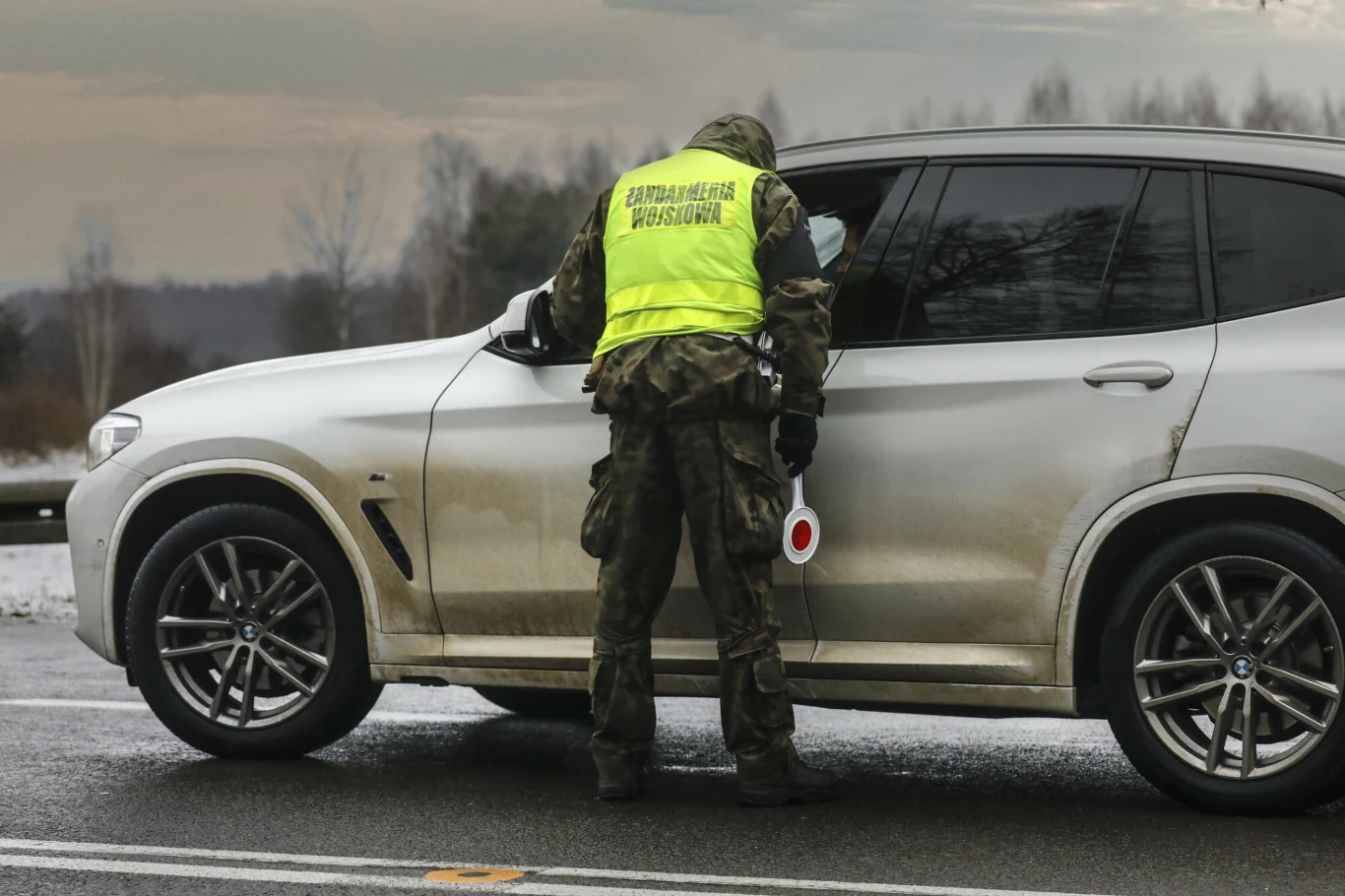 Nie wszystkie auta wjadą na teren jednostek wojskowych. Funkcjonariusz Straży Granicznej w żółtej kamizelce kontroluje kierowcę srebrnego samochodu na drodze w zimowych warunkach. W tle widoczne drzewa i szare niebo.