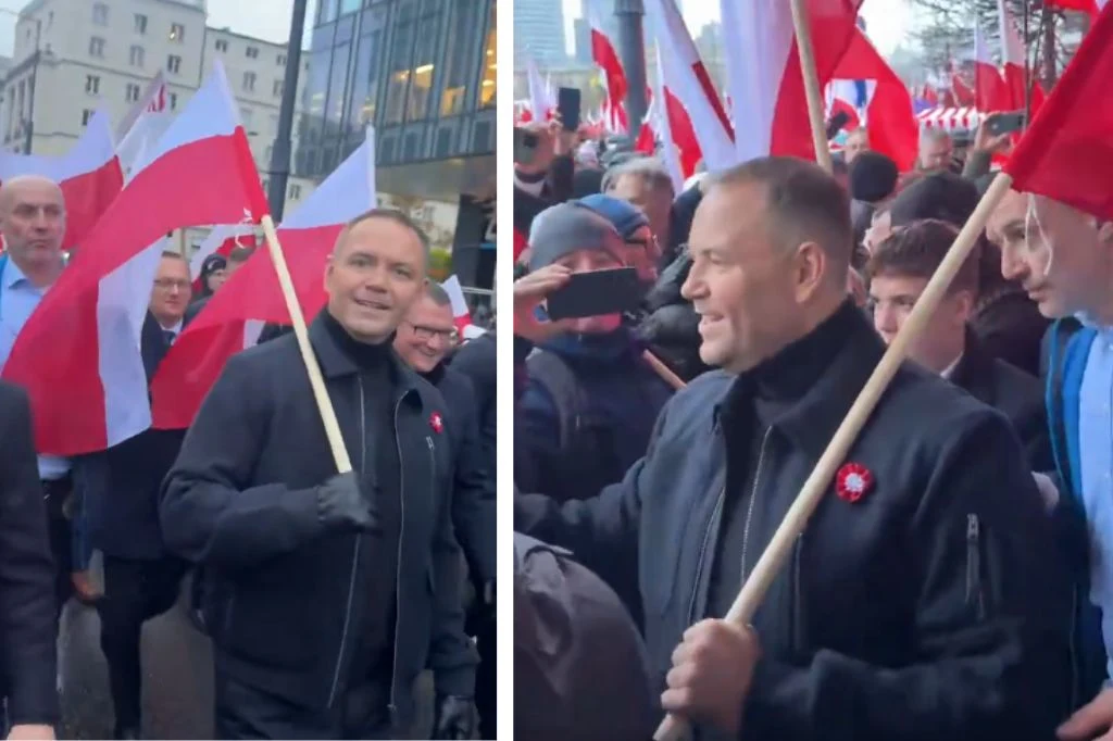 Karol Nawrocki podczas Marszu Niepodległości w Warszawie Tłum ludzi maszerujących ulicą z biało-czerwonymi flagami w rękach, w centrum znajduje się mężczyzna uśmiechający się i trzymający flagę, w tle widać wysokie budynki oraz innych uczestników pochodu.
