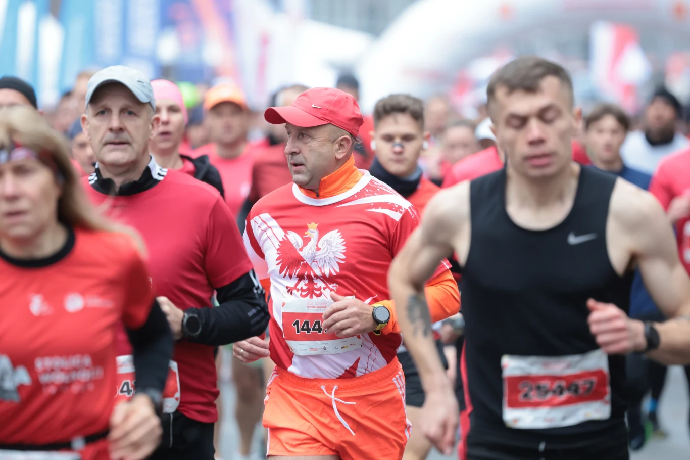 Grupa biegaczy w trakcie masowego biegu ulicznego, dominują czerwone stroje sportowe, jeden z zawodników ubrany w koszulkę z orłem na piersi; tło rozmyte, skupienie na twarzach i sylwetkach zawodników.