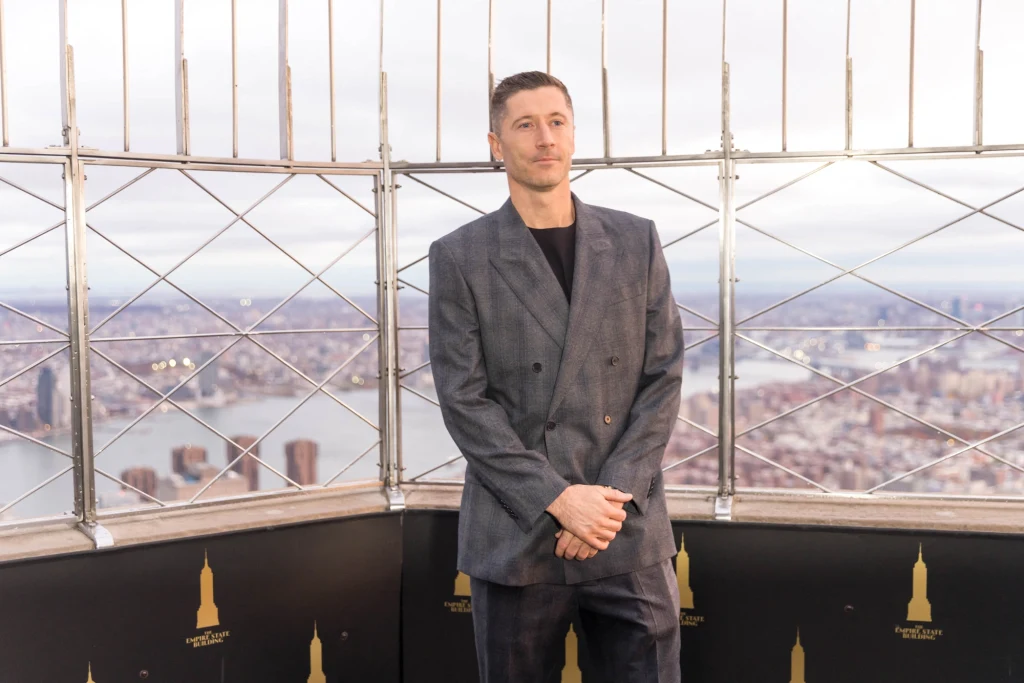 Robert Lewandowski Mężczyzna w szarym garniturze stoi na tarasie widokowym, w tle panorama miasta z rozpoznawalnymi wieżowcami oraz metalowa balustrada zabezpieczająca taras.