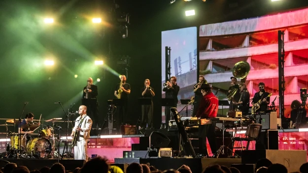 Zespół muzyczny występuje na scenie podczas koncertu, z licznymi instrumentami i sekcją dętą na podwyższeniu, kolorowe światła sceniczne i duży ekran z wizualizacjami stanowią tło, przed sceną zgromadzona jest publiczność.